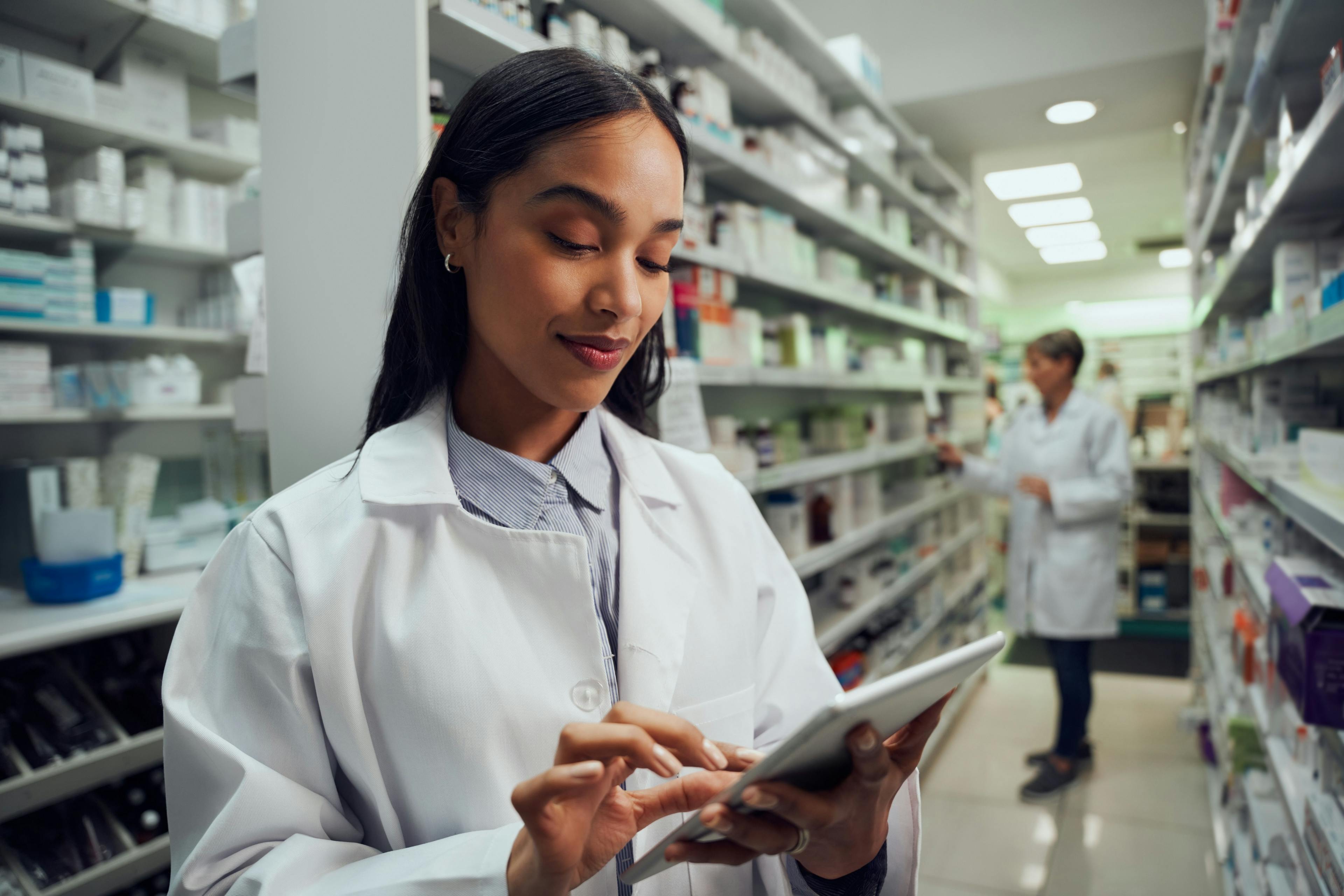 Smiling young female worker in pharmacy wearing labcoat checking inventory using digital tablet | Image Credit: StratfordProductions - stock.adobe.com