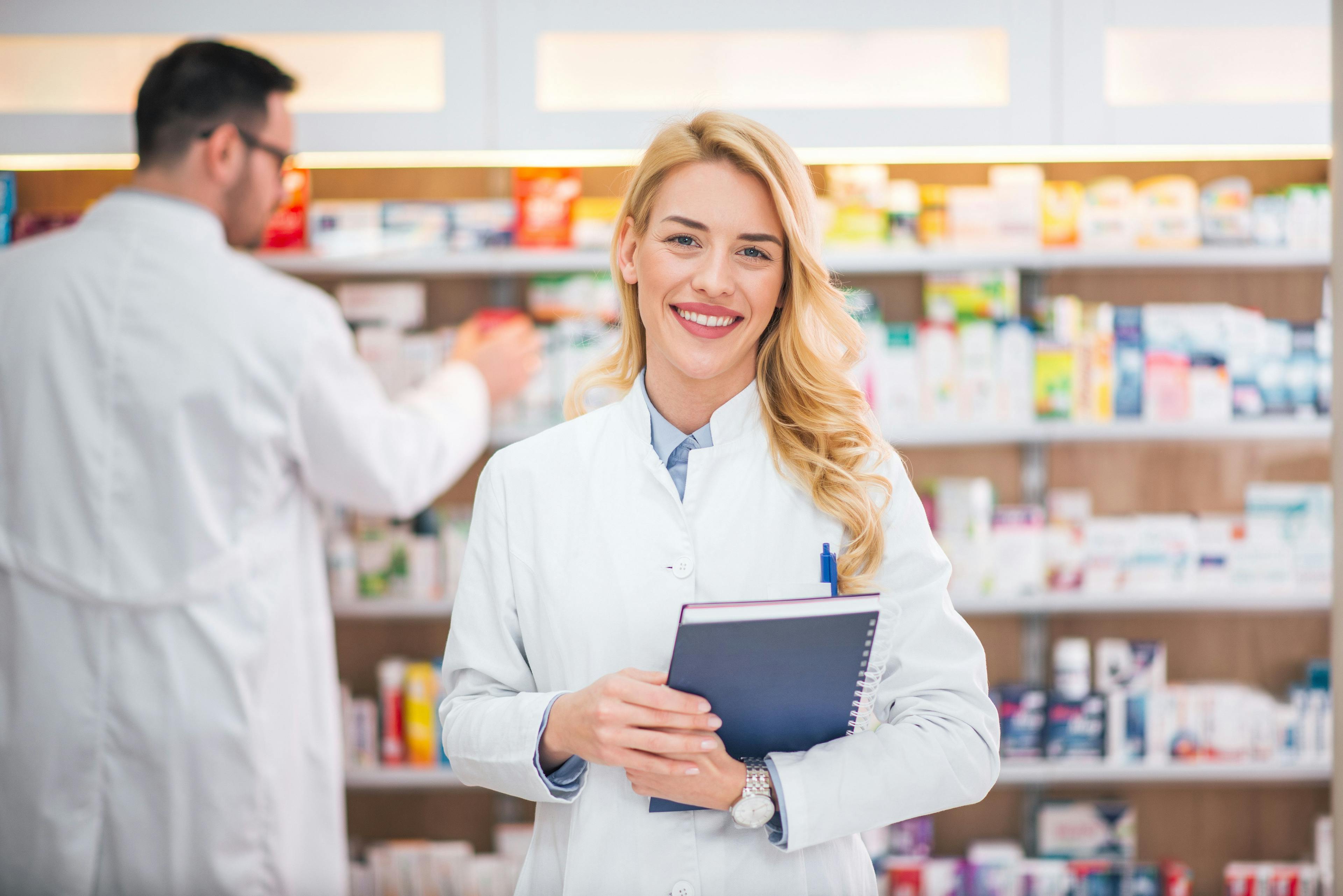 Portrait of a smiling female pharmacist, male colleague working with drugs in the background - Image credit: Bnenin | stock.adobe.com