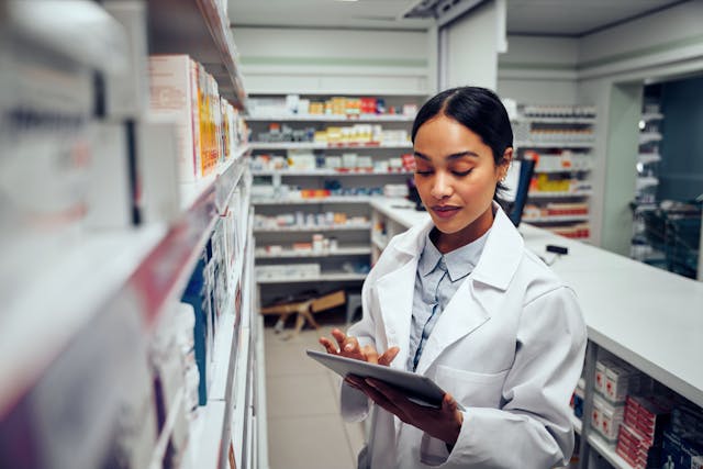 Young female pharmacist checking inventory of medicines in pharmacy using digital tablet wearing labcoat standing behind counter | Image Credit: StratfordProductions - stock.adobe.com