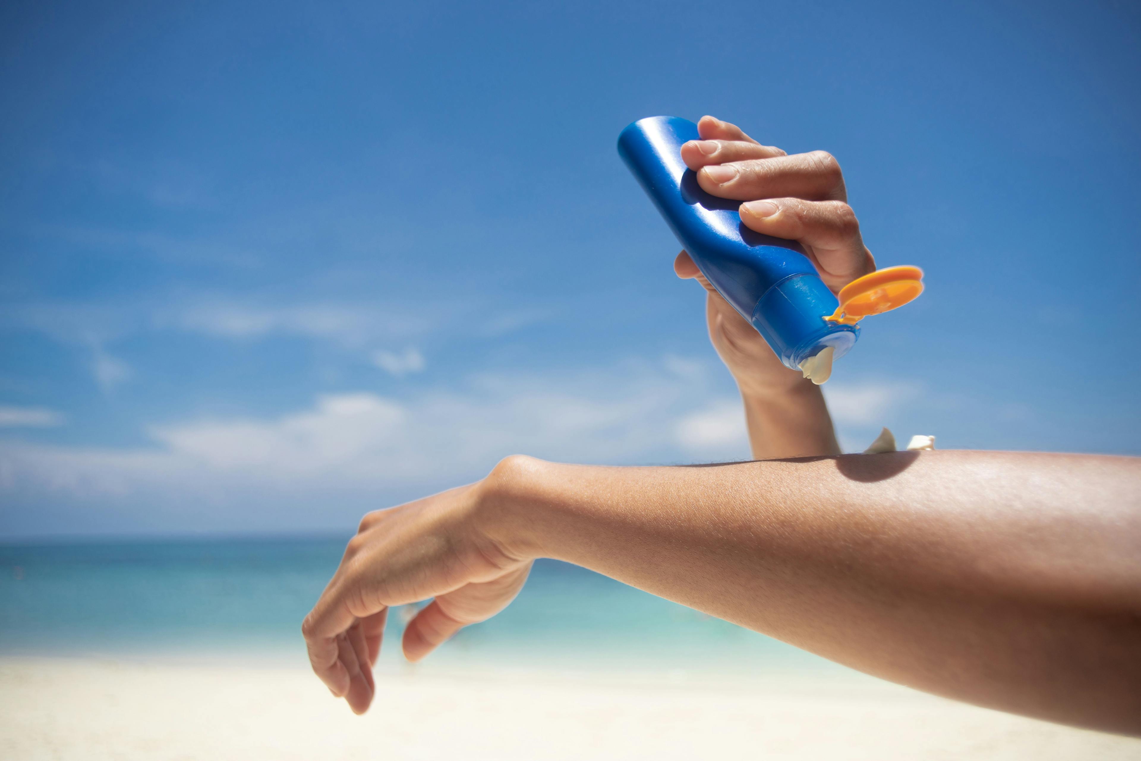 Woman applying sunscreen on her hands from a bottle on the beach with the sea in the background | Image Credit: lesterman - stock.adobe.com