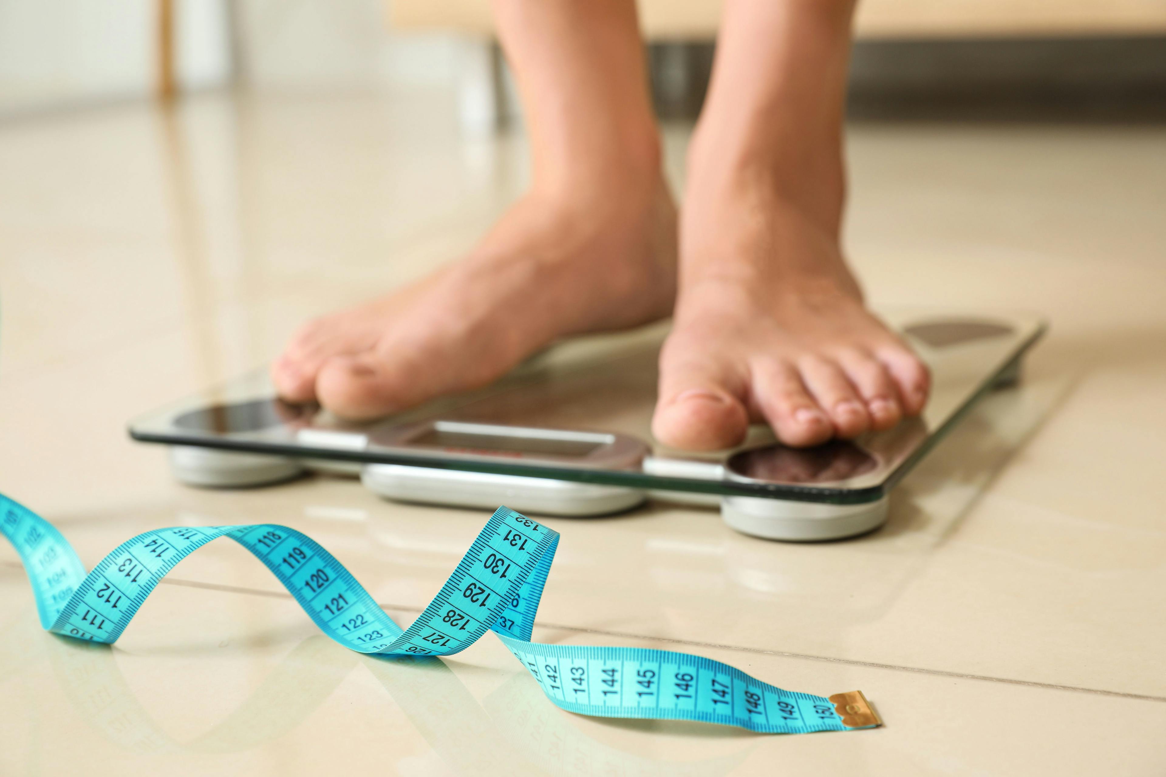 Tape in front of woman standing on floor scales indoors. Obesity | Image Credit: New Africa - stock.adobe.com