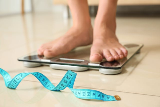 Tape in front of woman standing on floor scales indoors. Obesity | Image Credit: New Africa - stock.adobe.com