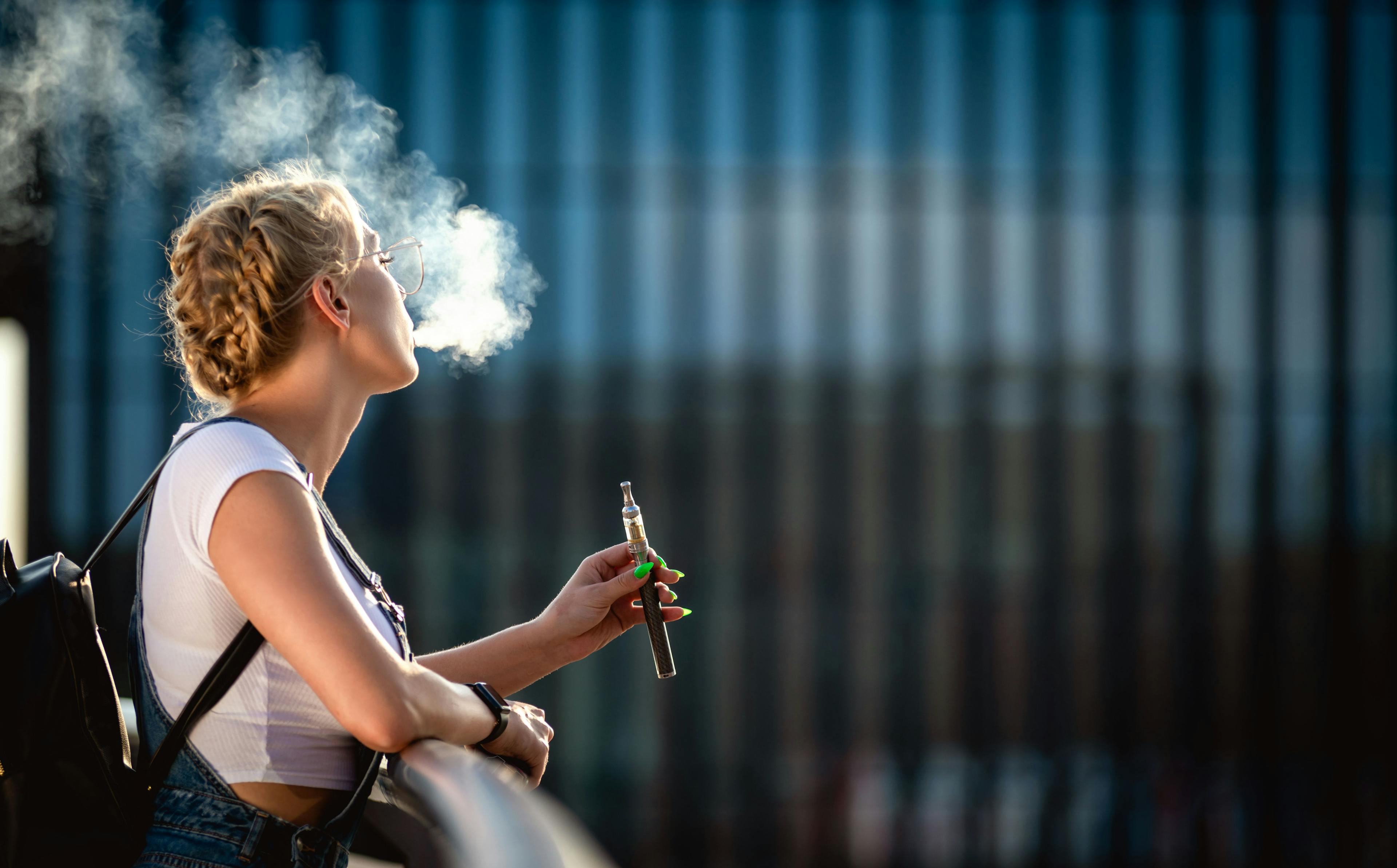 Young woman outdoor vaping e-cigarette on modern city buildings background | Image Credit: leszekglasner - stock.adobe.com
