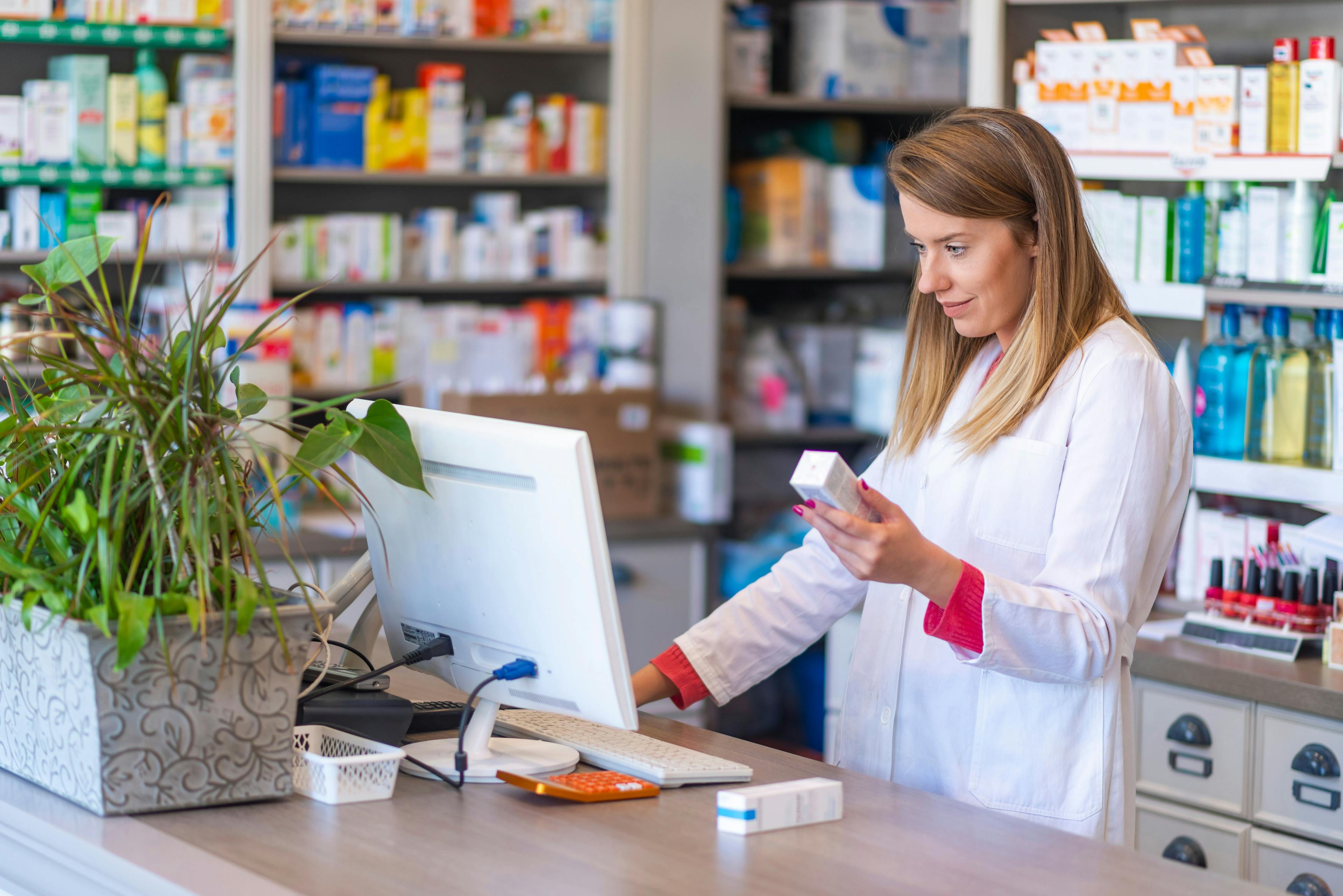 Portrait of young female pharmacist holding medication while using computer at pharmacy counter | Image Credit: Dragana Gordic - stock.adobe.com