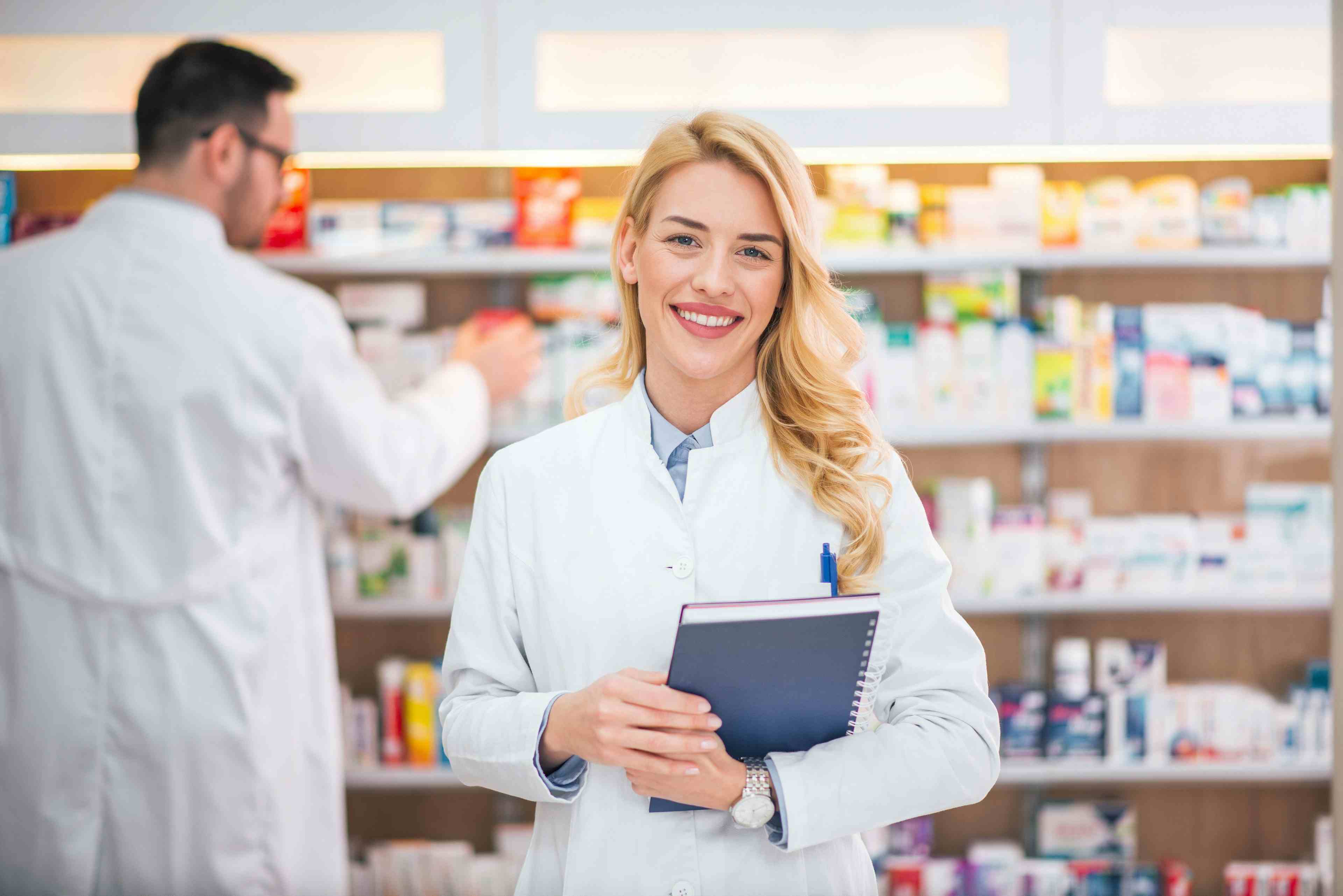 Portrait of a smiling female pharmacist, male colleague working with drugs in the background. | Image Credit: bnenin - stock.adobe.com