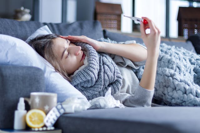 Young woman with fever and headache is measuring temperature with thermometer, treated at home. Winter cold and flu concept. | Image Credit: Monstar Studio - stock.adobe.com