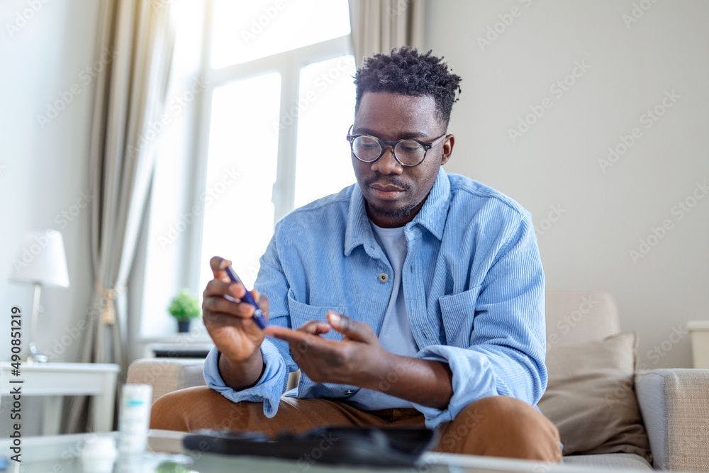 African man is sitting at the sofa and taking blood from his finger due to diabetes. The daily life of a man of African-American ethnicity person with a chronic illness who is using glucose tester. | Image Credit: Graphicroyalty - stock.adobe.com