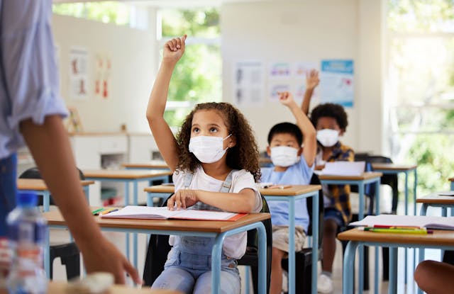 Covid, education and learning of eager, smart and clever children raising hands and wearing masks in a classroom. Teacher or educator asking questions for knowledge test at kids school in a pandemic | Image credit: Nina/peopleimages.com - stock.adobe.com
