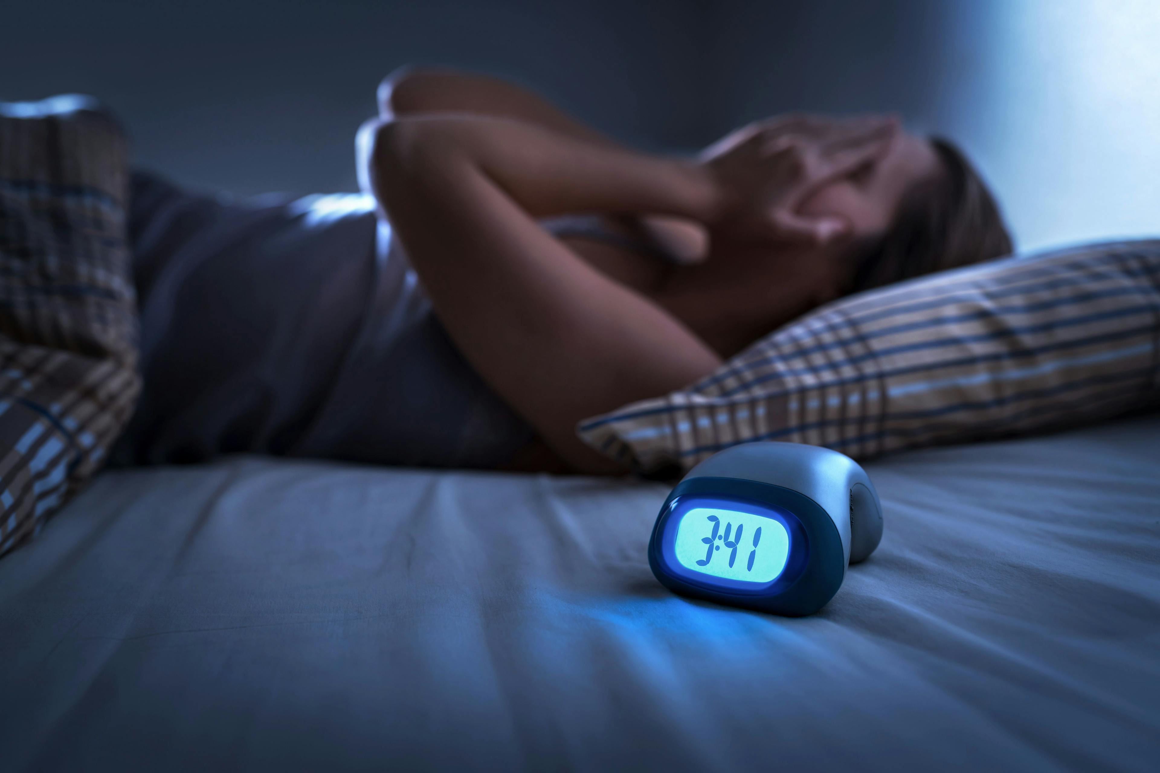 Sleepless woman suffering from insomnia, sleep apnea or stress. Tired and exhausted lady. Headache or migraine. Awake in the middle of the night. Frustrated person with problem. Alarm clock with time | Image Credit: terovesalainen - stock.adobe.com
