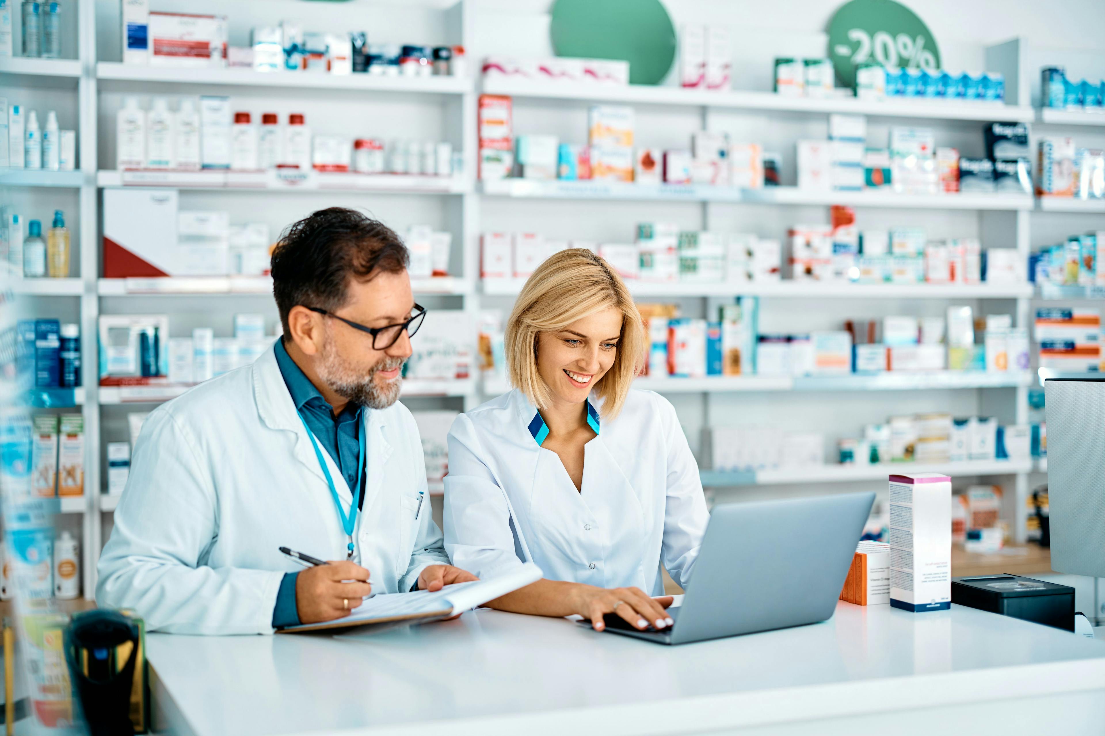 Happy pharmacist using laptop while her colleagues is taking notes in pharmacy. | Image Credit: Drazen - stock.adobe.com